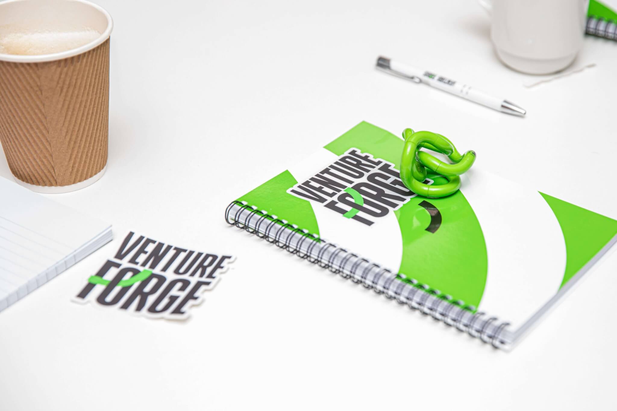Venture Forge branded notebook, sticker, and pen on a desk with coffee cup.